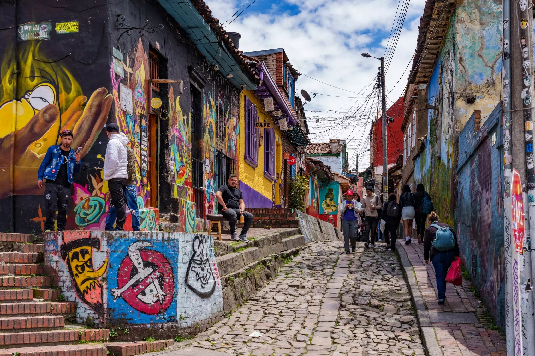 People are seen on a narrow cobbled street in a city. Vibrant murals and street art covers the walls of buildings along the street.
