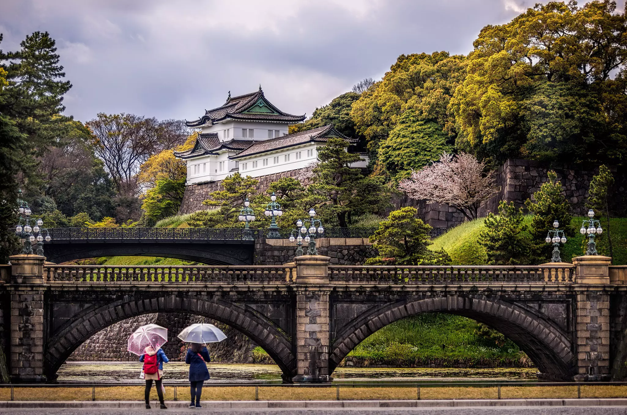 Two women stood beneath umbrellas stand in front of a bridge and a hilltop palace
