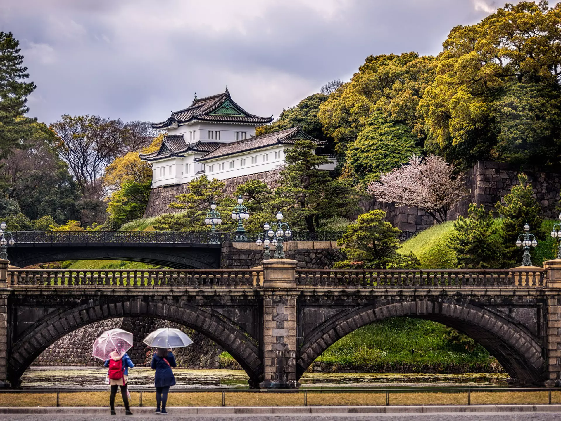 Two women stood beneath umbrellas stand in front of a bridge and a hilltop palace