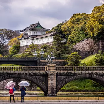 Two women stood beneath umbrellas stand in front of a bridge and a hilltop palace