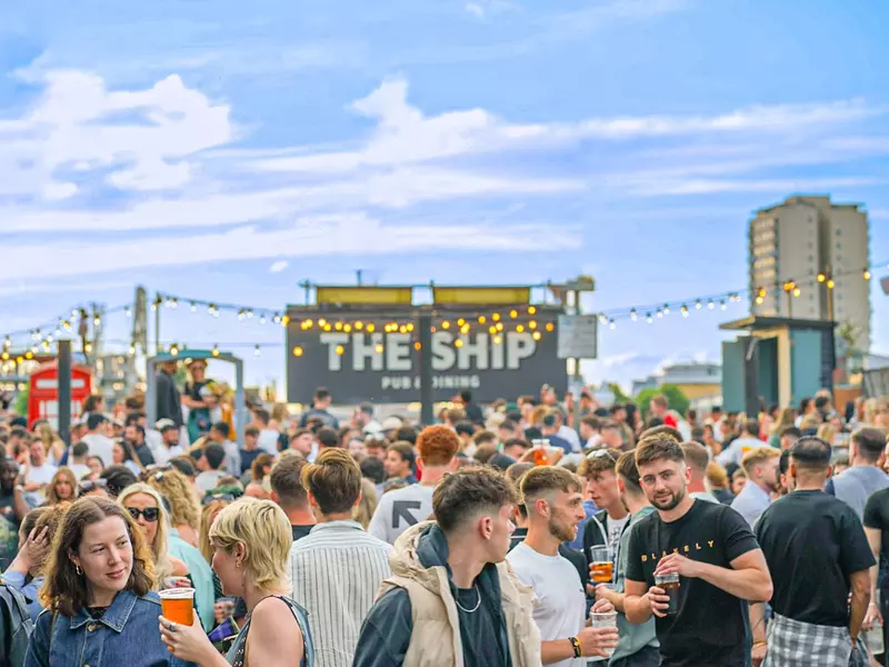 A crowd of people at an outdoor pub with a large sign with its logo, "The Ship"