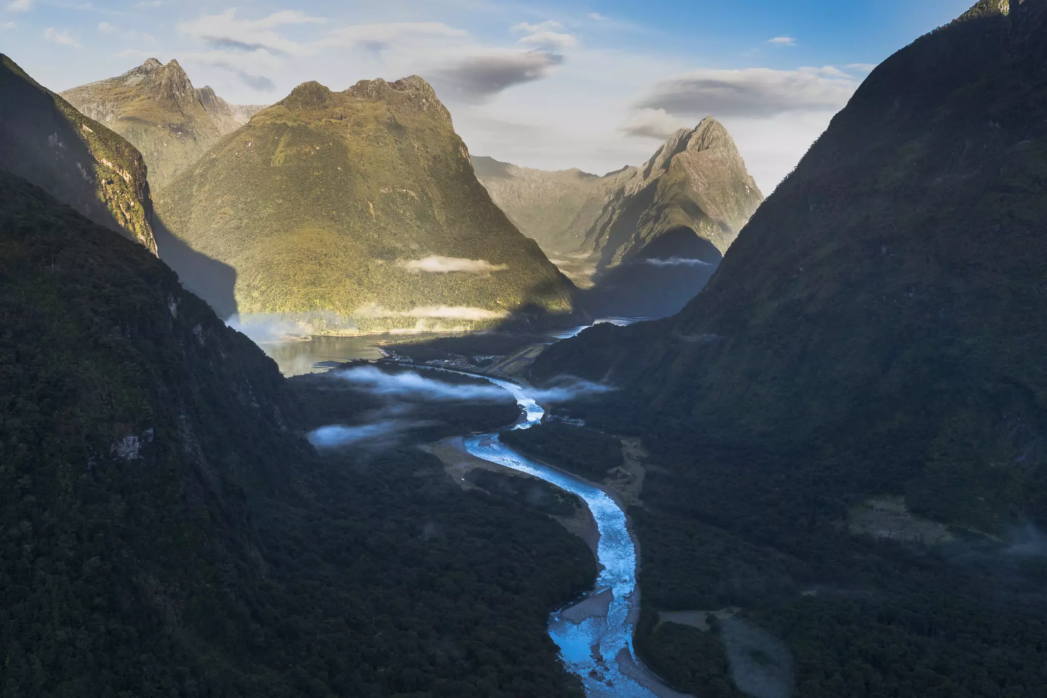 An aerial view of a mountainous landscape with a bright blue stream cutting through it; there is a sharp contrast on the hills between sunlight and shadow.
