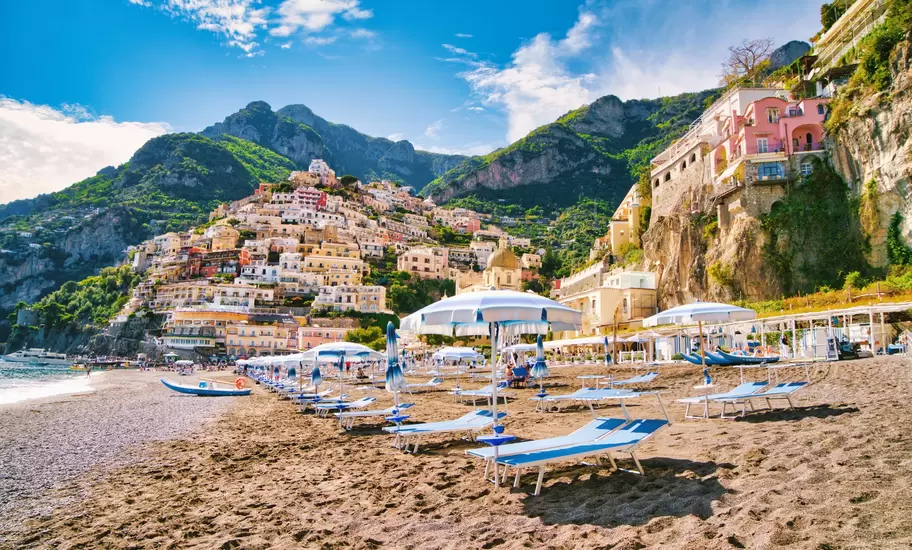 Rows of sun loungers and umbrellas on a dark sand beach below pastel houses stacked up a steep hillside