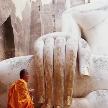 Monk in an orange robe kneeling beside the huge stone hand of a seated white Buddha at Wat Si Chum, Sukhothai, Thailand.