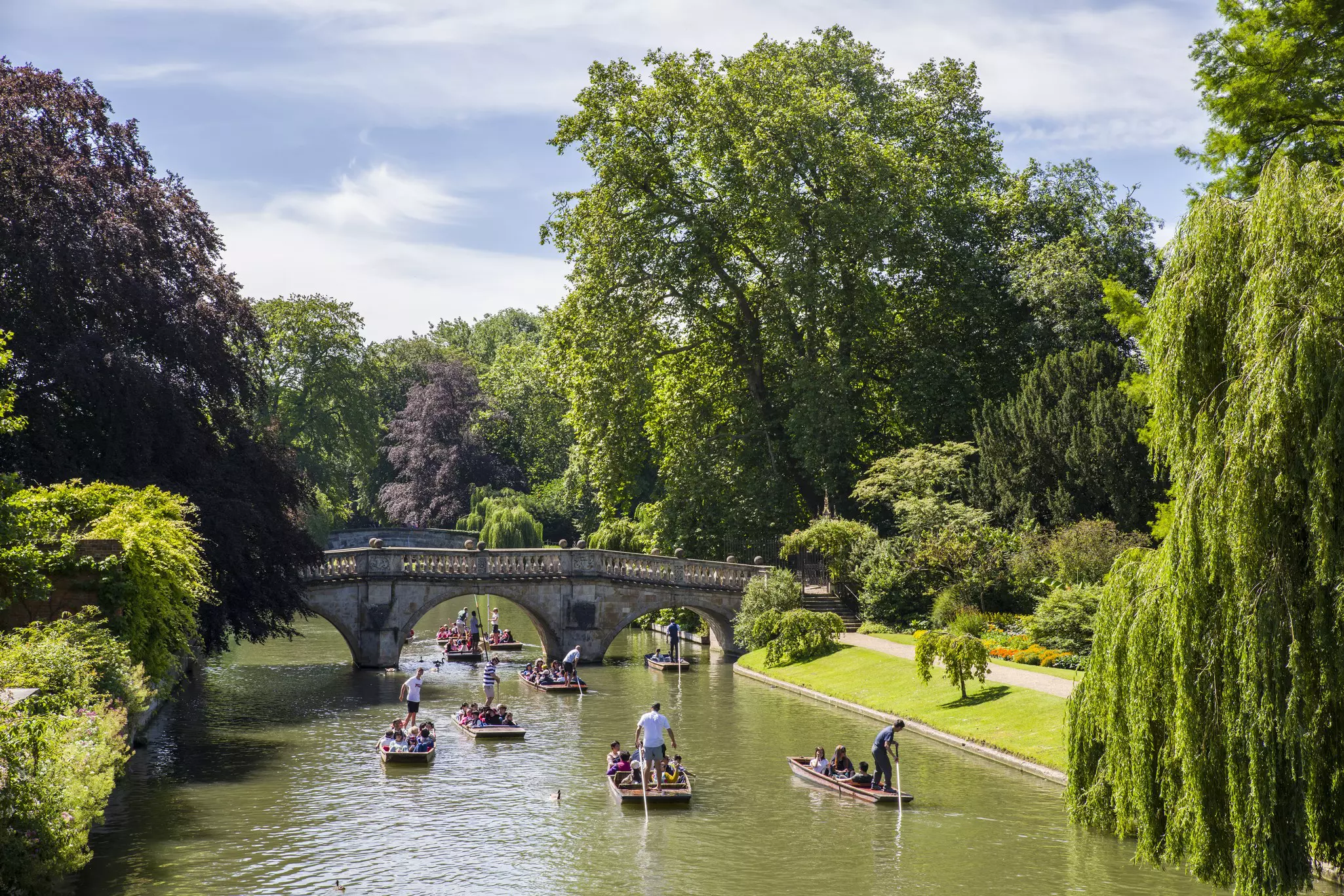 A stone bridge crosses a greenish river where several small boats are filled with people; trees and greenery are on both shores.