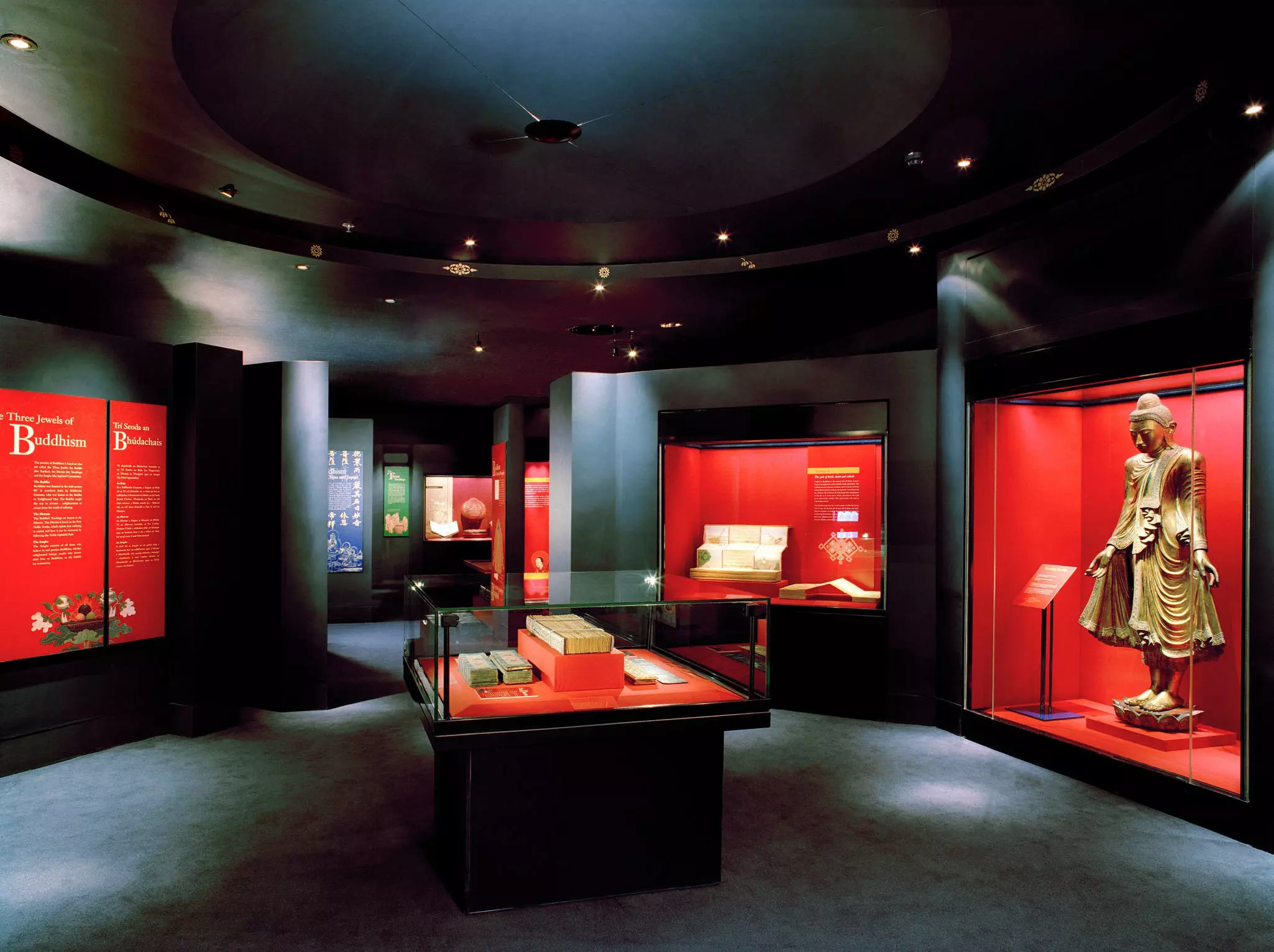 A series of books and a large Buddha statue in display cases in a dark gallery.