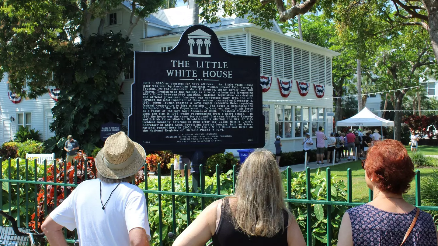 People read a historic marker by an iron fence in front of a white-painted house in a tropical location.