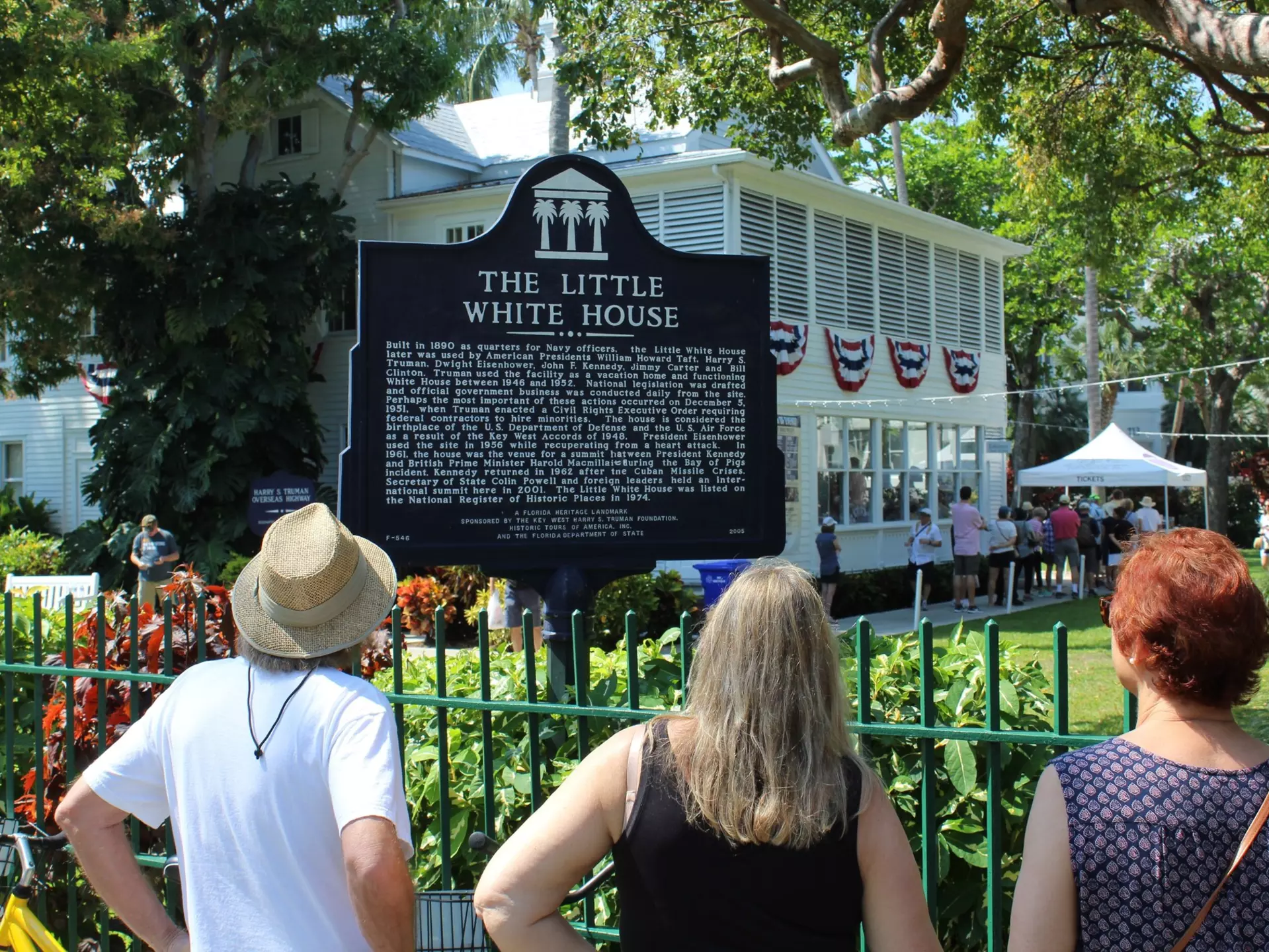 People read a historic marker by an iron fence in front of a white-painted house in a tropical location.