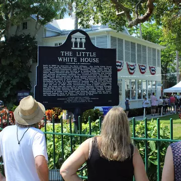 People read a historic marker by an iron fence in front of a white-painted house in a tropical location.