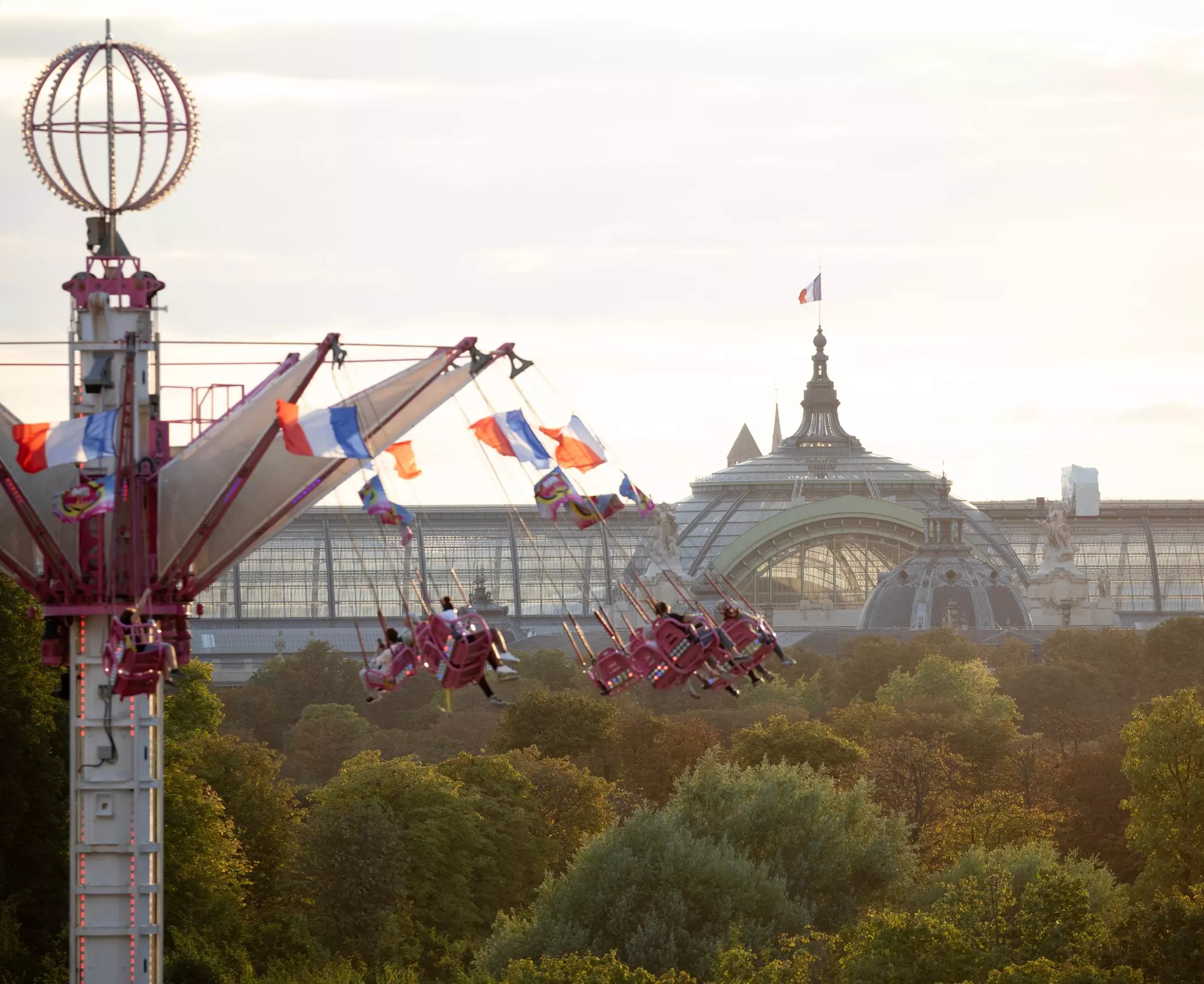 Carousel of flying swings spins over green trees with Grand Palais in Paris in the background