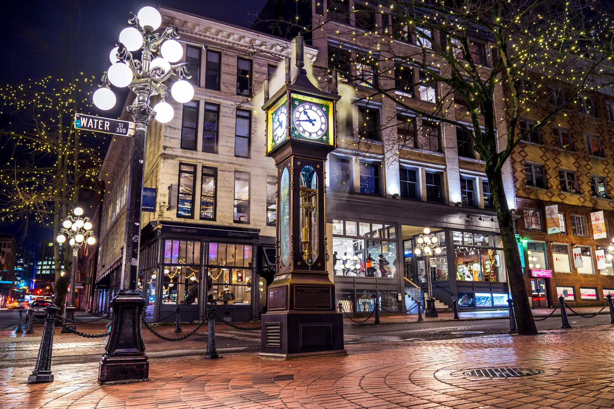 A clock in the center of a neighborhood with a shop-lined street.