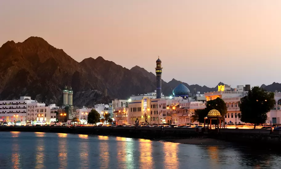 Muscat's Mutrah Corniche at dusk, with a blue-domed mosque, minarets and lit waterfront below rugged mountains, Oman