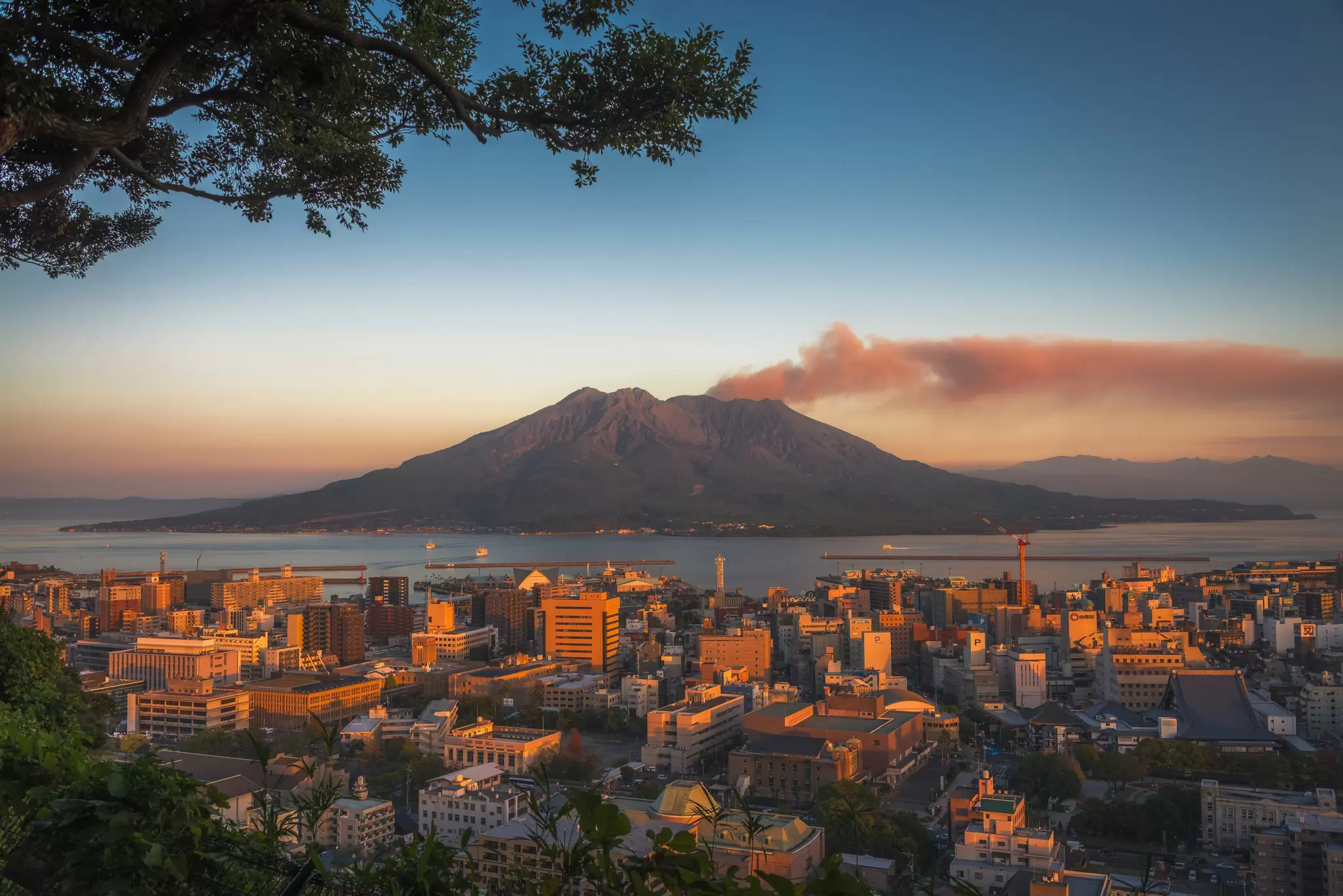 Sunset over a bay city skyline with a smoking volcano releasing an ash plume across the sky