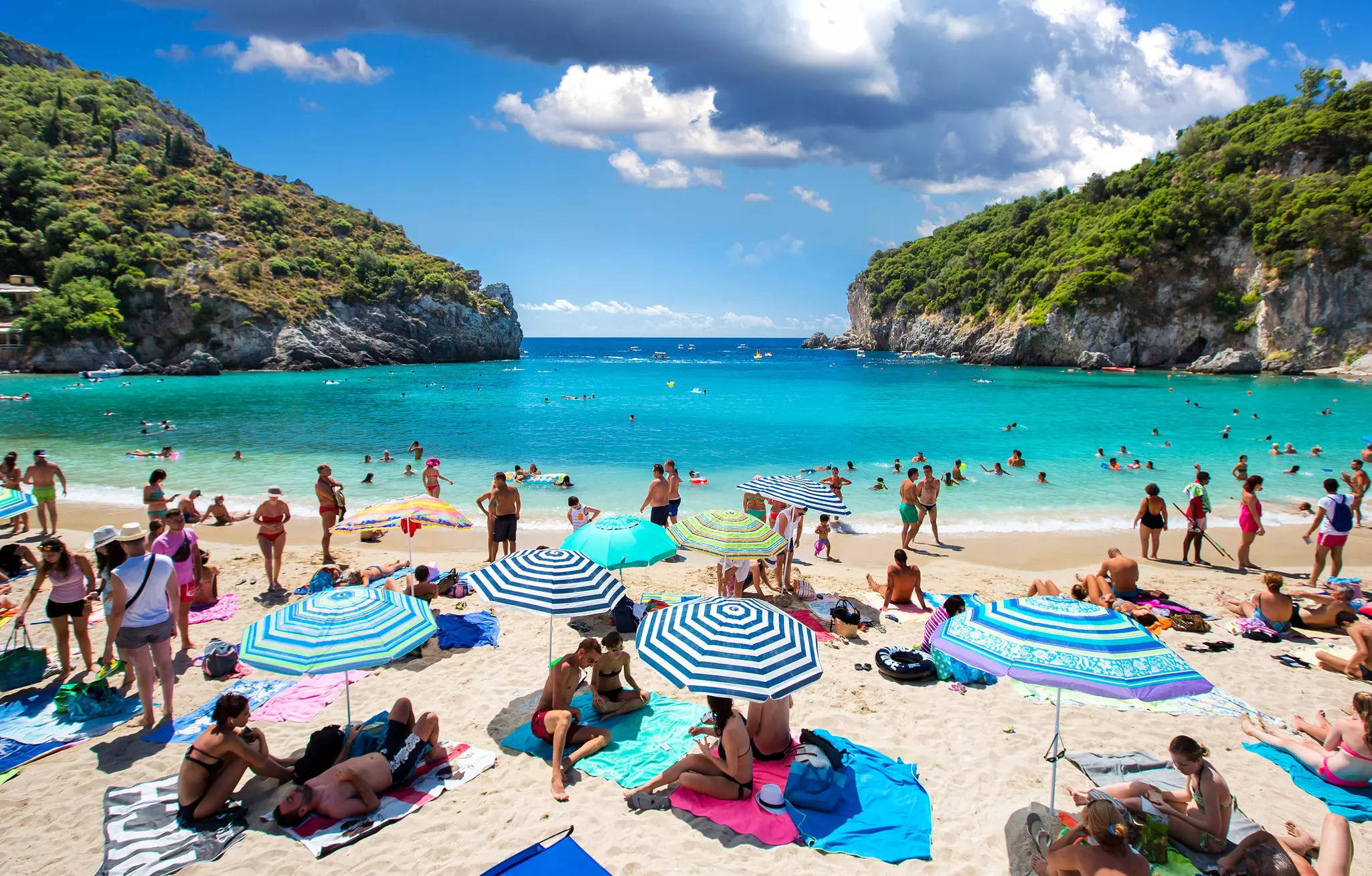 A small beach in a cove is crowded with people lying on towels and under umbrellas.