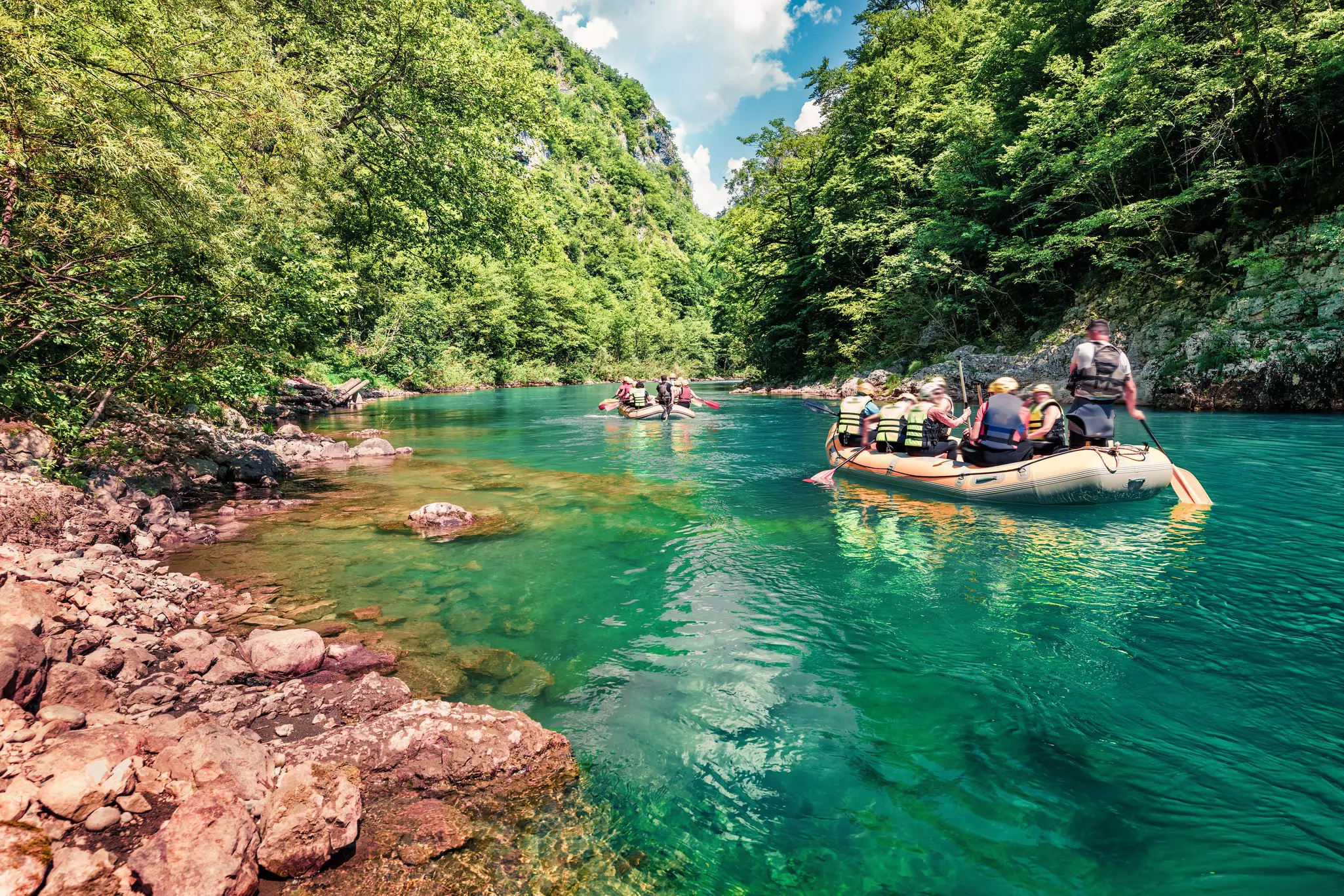 Two inflatable boats fully loaded with passengers in life vests make their way along a river near banks covered in greenery