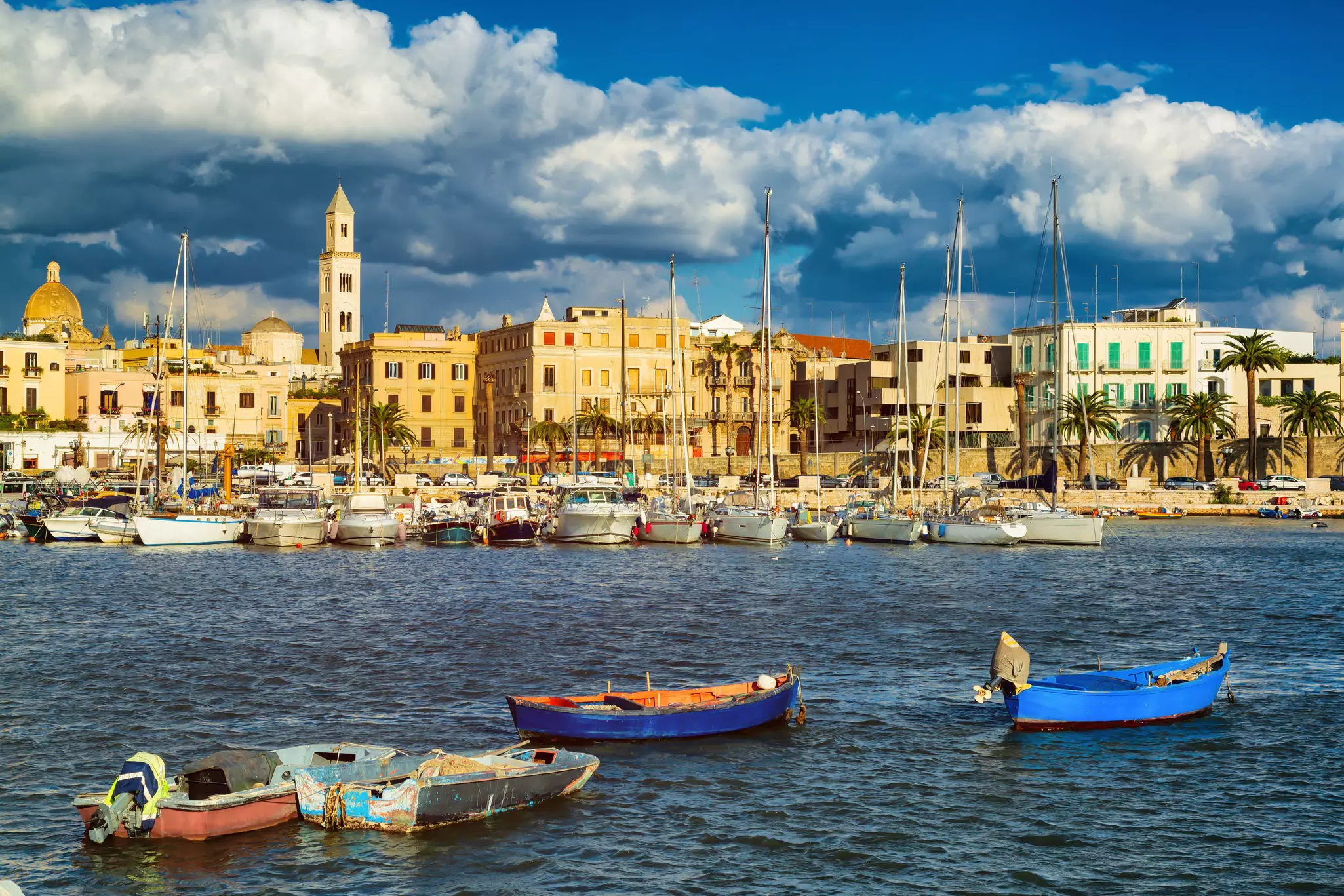 Boats are moored in a harbor in a city. Stone buildings line the waterfront.