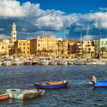 Boats are moored in a harbor in a city. Stone buildings line the waterfront.