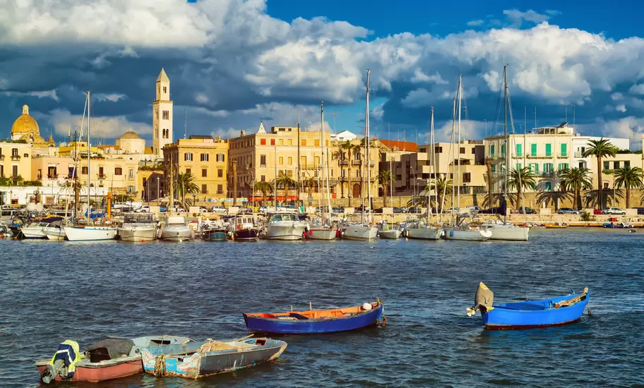 Boats are moored in a harbor in a city. Stone buildings line the waterfront.