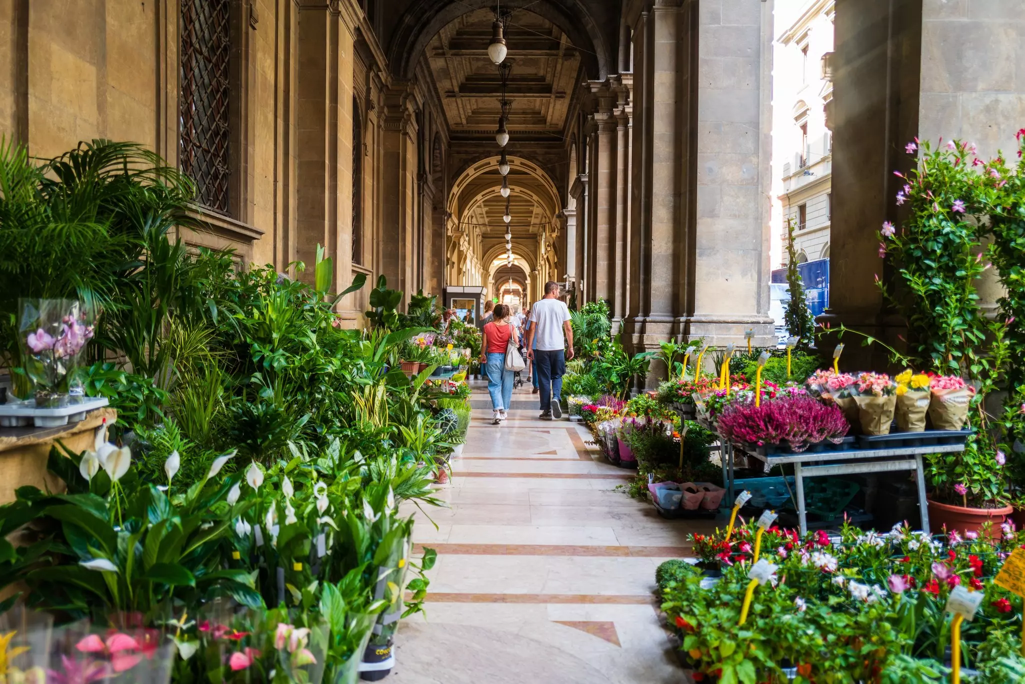People walk through a plant market under stone archways.