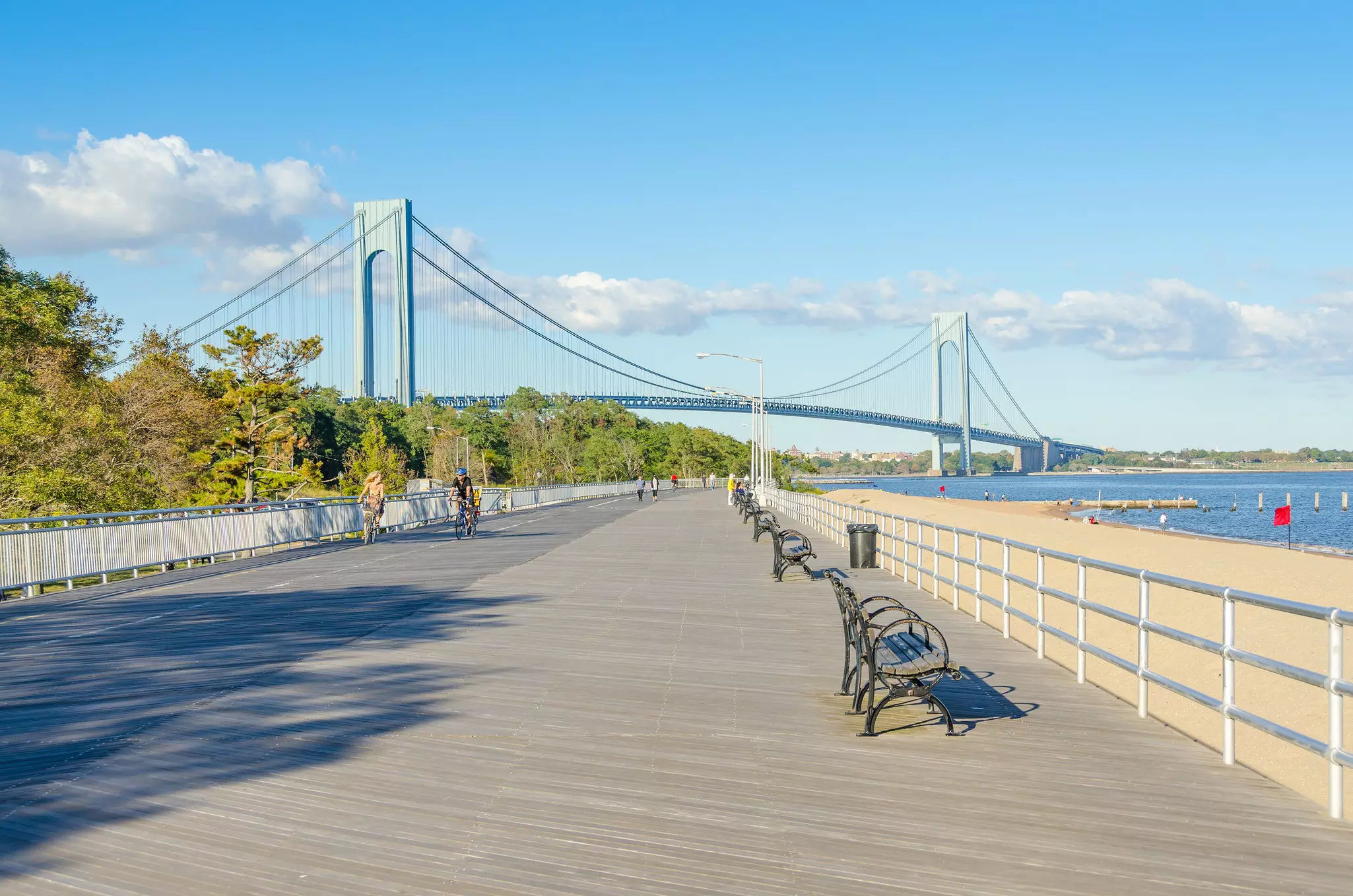 Two cyclists pedal along a beach-side boardwalk away from a large suspension bridge in the distance.