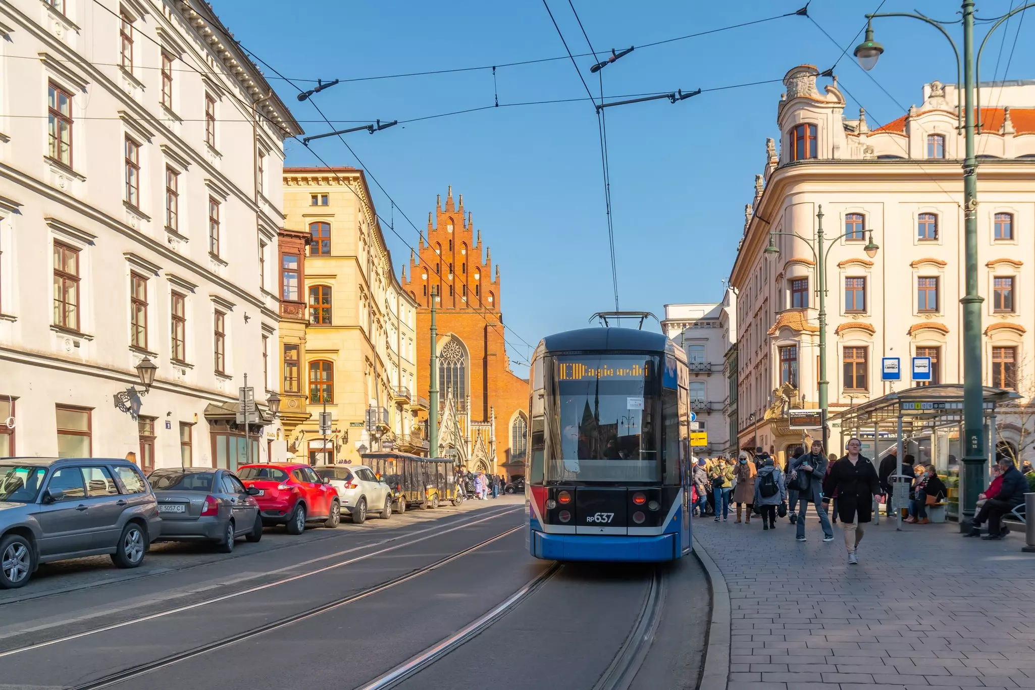 A blue tram passing a stop for passengers in Kraków; a church is visible at the end of the roadway.