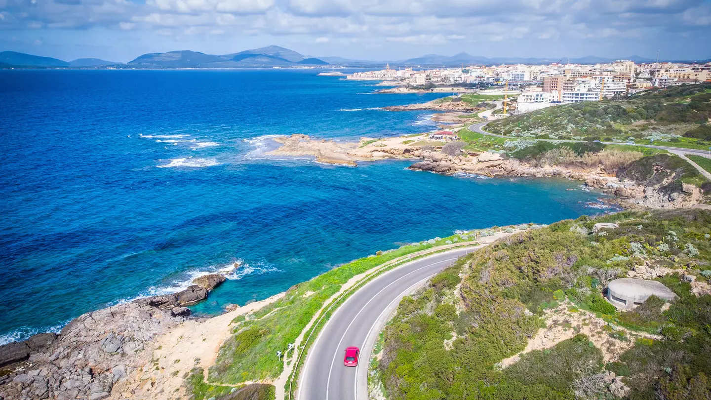 An aerial view of a red car driving on a road along a coastal road. A town is seen in the distance.