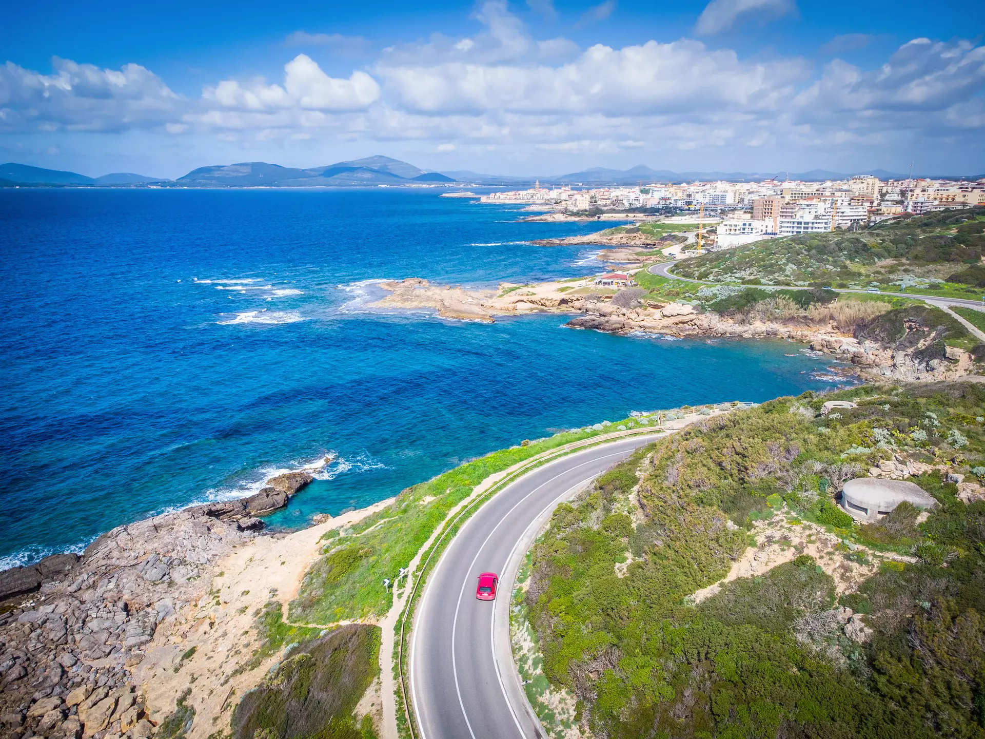 An aerial view of a red car driving on a road along a coastal road. A town is seen in the distance.