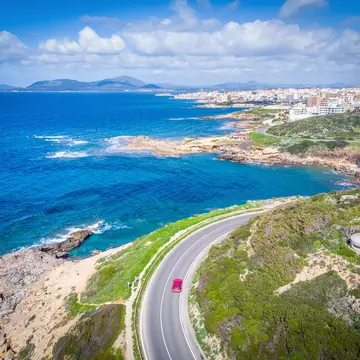 An aerial view of a red car driving on a road along a coastal road. A town is seen in the distance.
