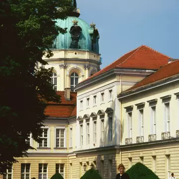 Pale yellow baroque palace with a green copper dome and gold statue on top, a group of young people on a bench in front.