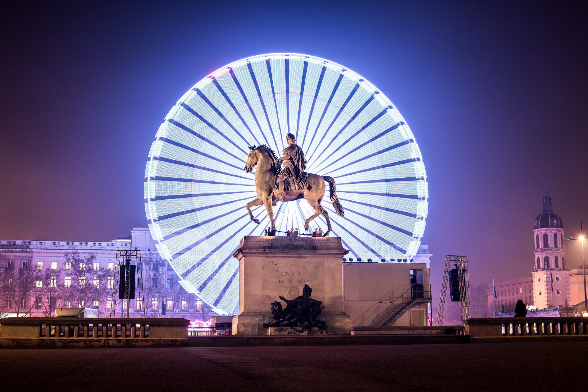 Big illuminated ferris wheel behind the Statue of Louis XIV at night in the Place Bellecour area during the Festival of Lights in Lyon, France.