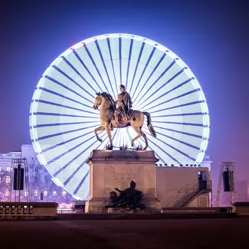 Big illuminated ferris wheel behind the Statue of Louis XIV at night in the Place Bellecour area during the Festival of Lights in Lyon, France.