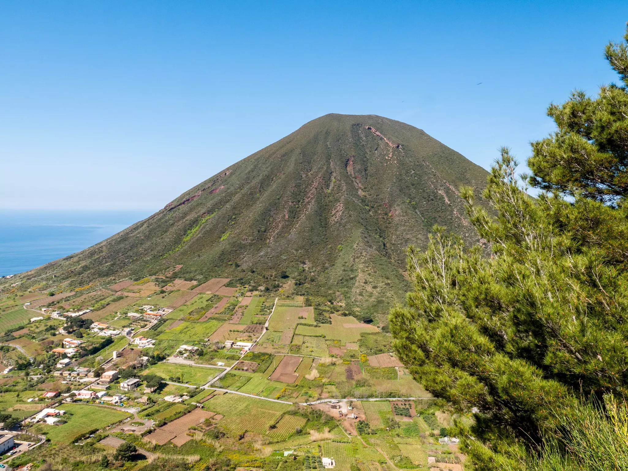 A vast coastal volcano with fertile land and crops growing on its side.
