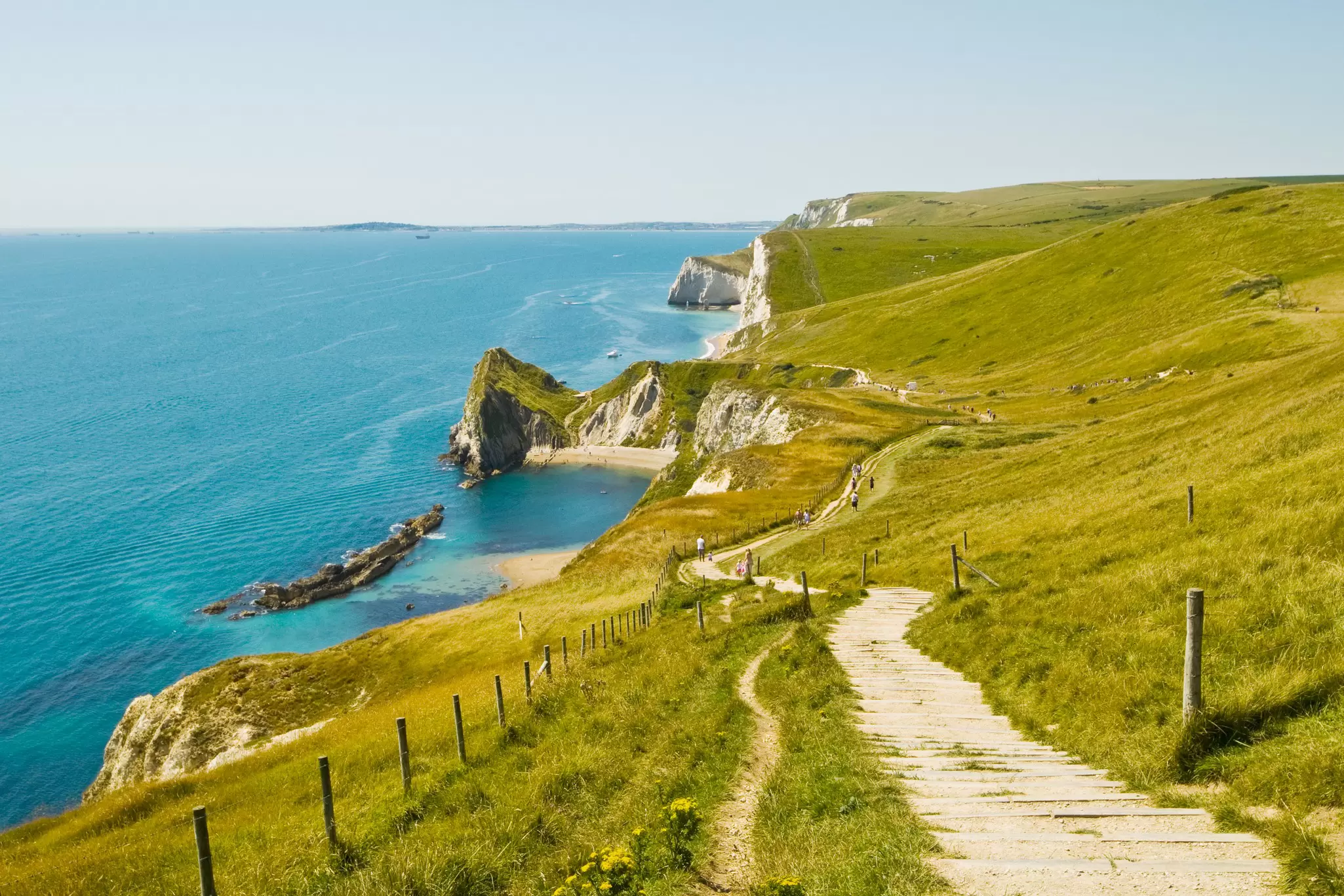 Hikers follow an undulating chalk path over white cliffs with sandy beaches below. 