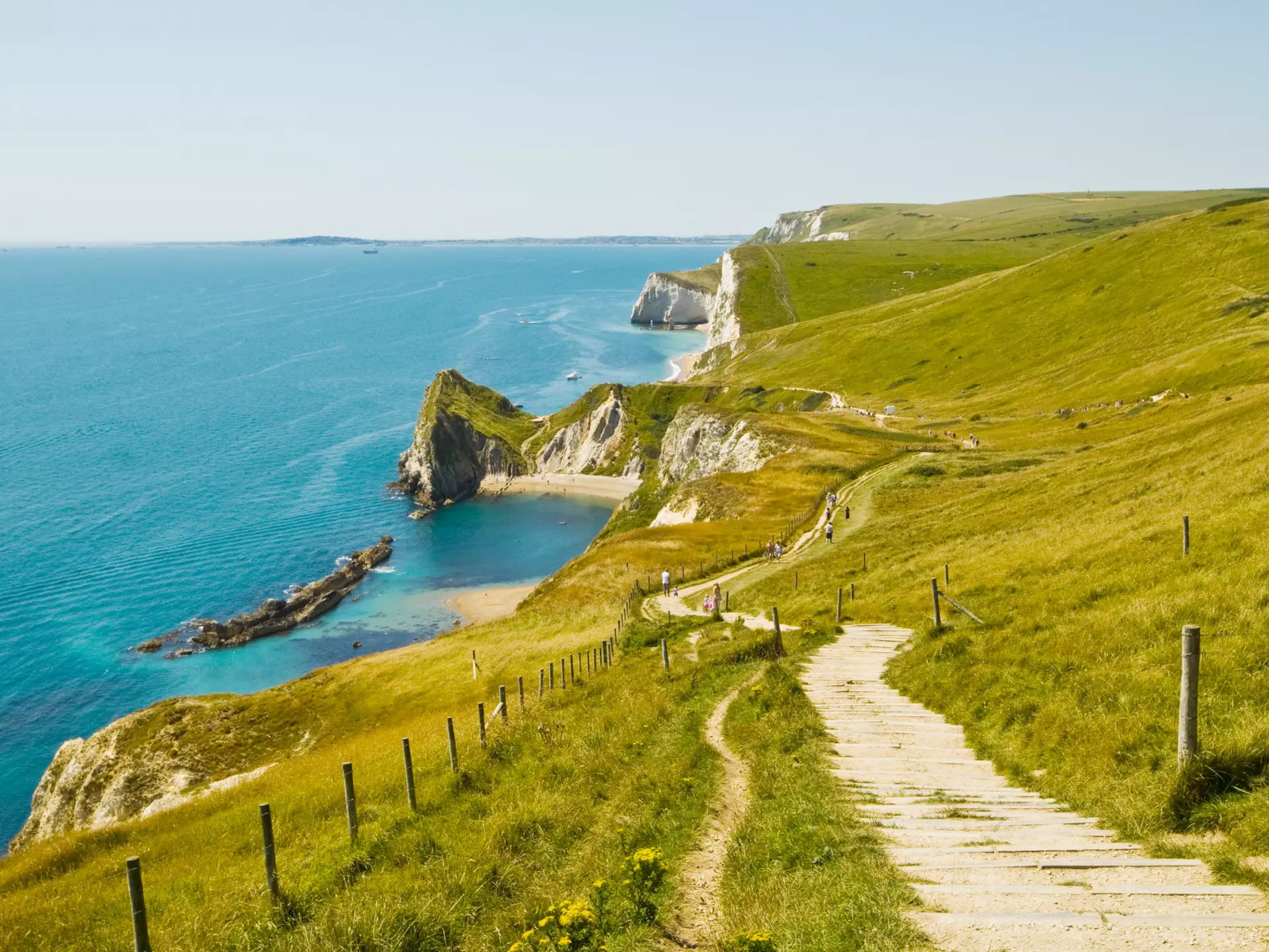 Hikers follow an undulating chalk path over white cliffs with sandy beaches below. 