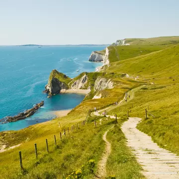 Hikers follow an undulating chalk path over white cliffs with sandy beaches below. 