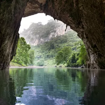 Still green water flowing out of a large cave mouth toward jungle-covered limestone cliffs under a misty sky.