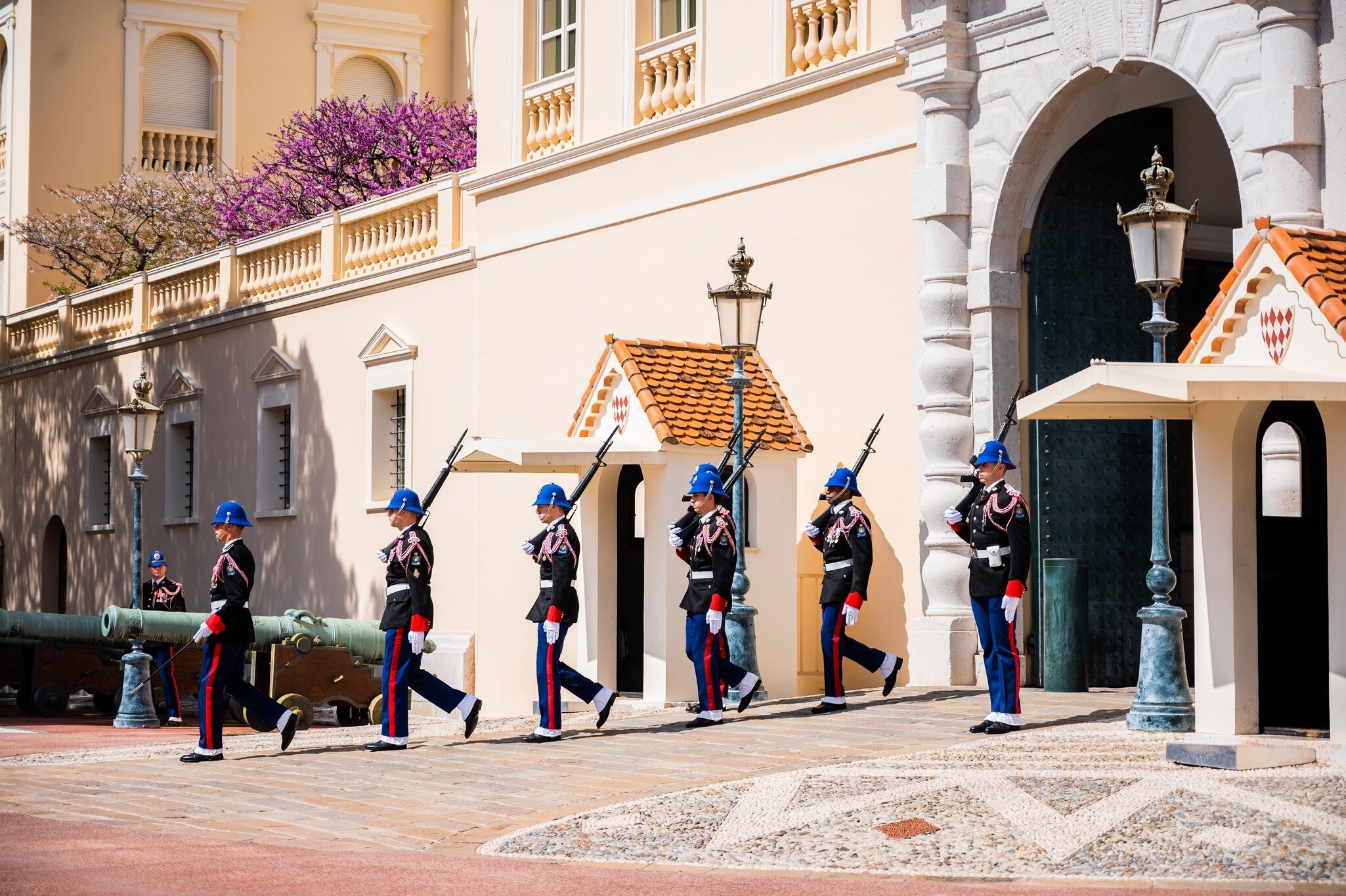 Six guards in dark tunics, blue helmets and red-striped trousers marching in file past sentry boxes and old bronze cannons outside a pale palace.