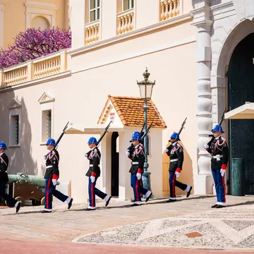 Six guards in dark tunics, blue helmets and red-striped trousers marching in file past sentry boxes and old bronze cannons outside a pale palace.