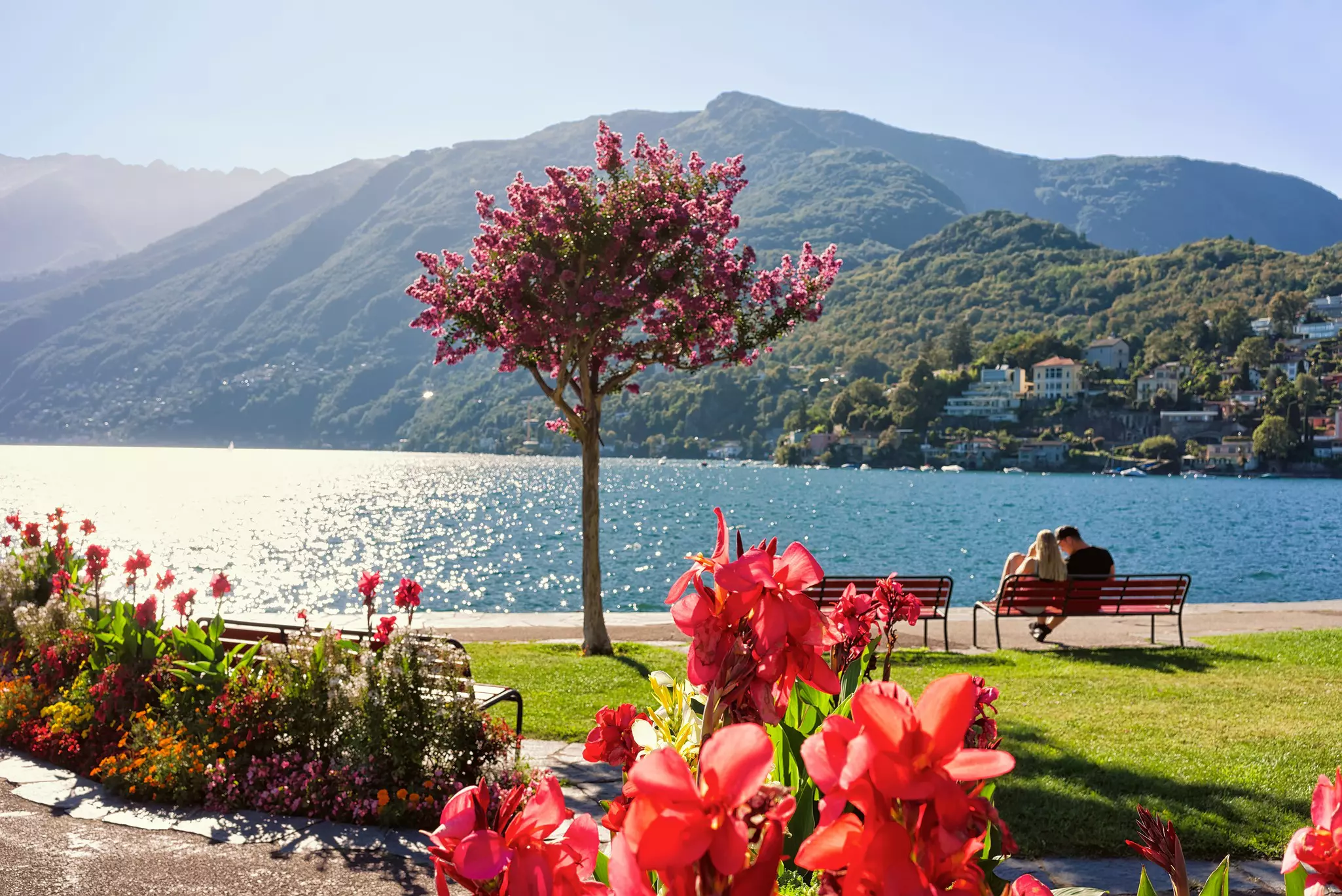 A couple sit on a bench on the promenade beside a lake glistening in the sunshine.