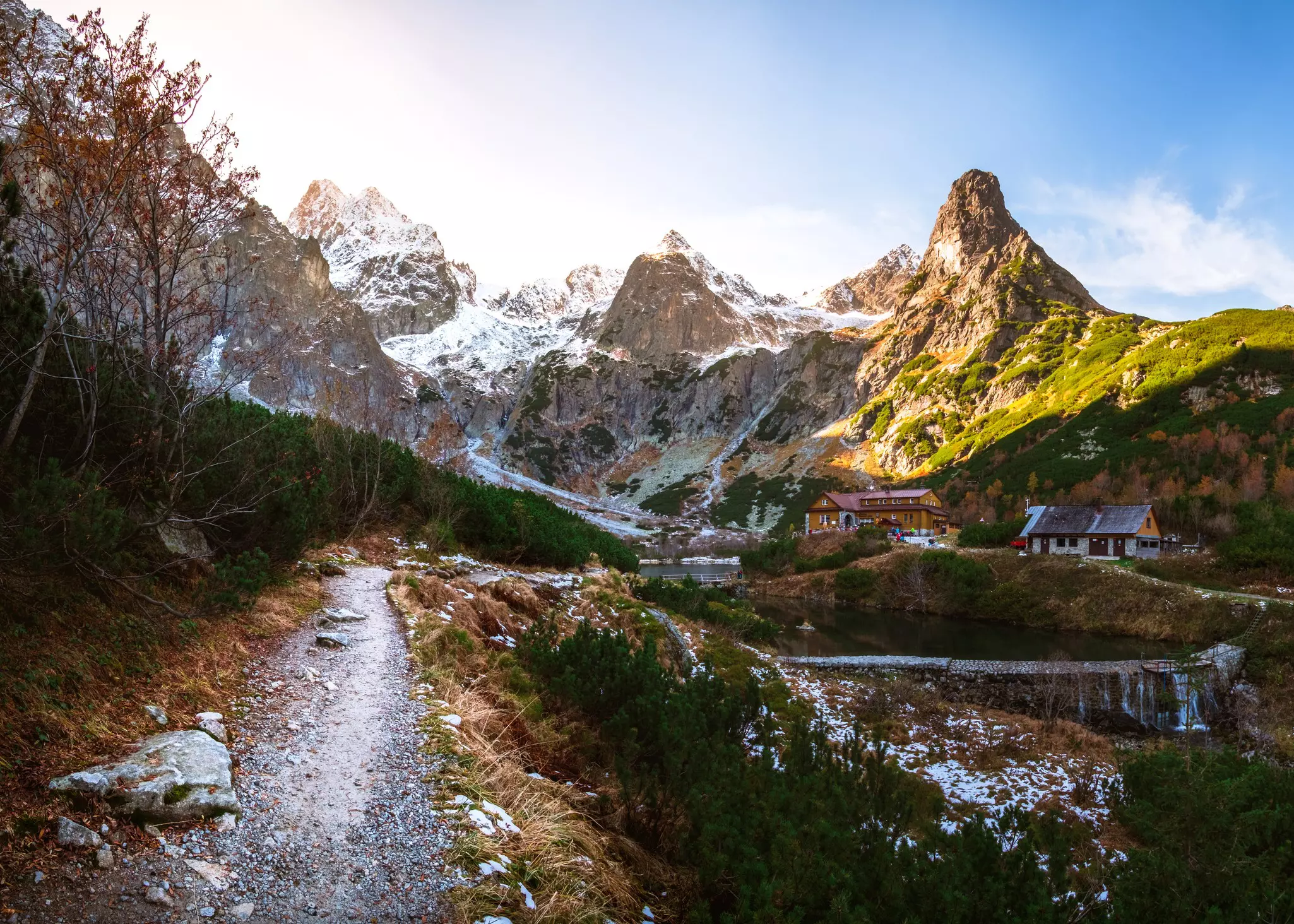 A path leads by a mountain lake. Light snow covers the ground, and sunlight hits snow-covered crags in the distance.