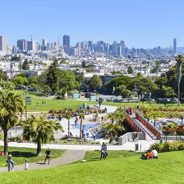 People relaxing on grassy slopes among palm trees in Dolores Park, San Francisco, with downtown high-rises on the horizon.