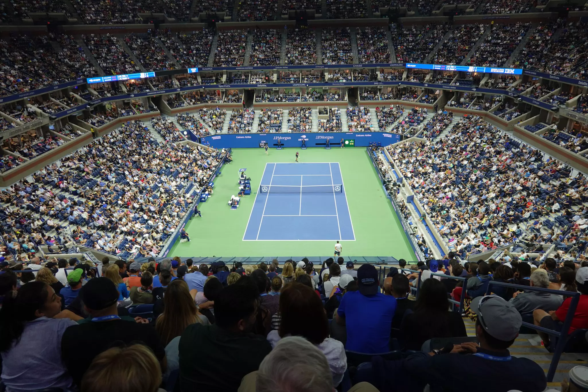 A crowded audiotorium with seats full of people watching a tennis match on the court below.
