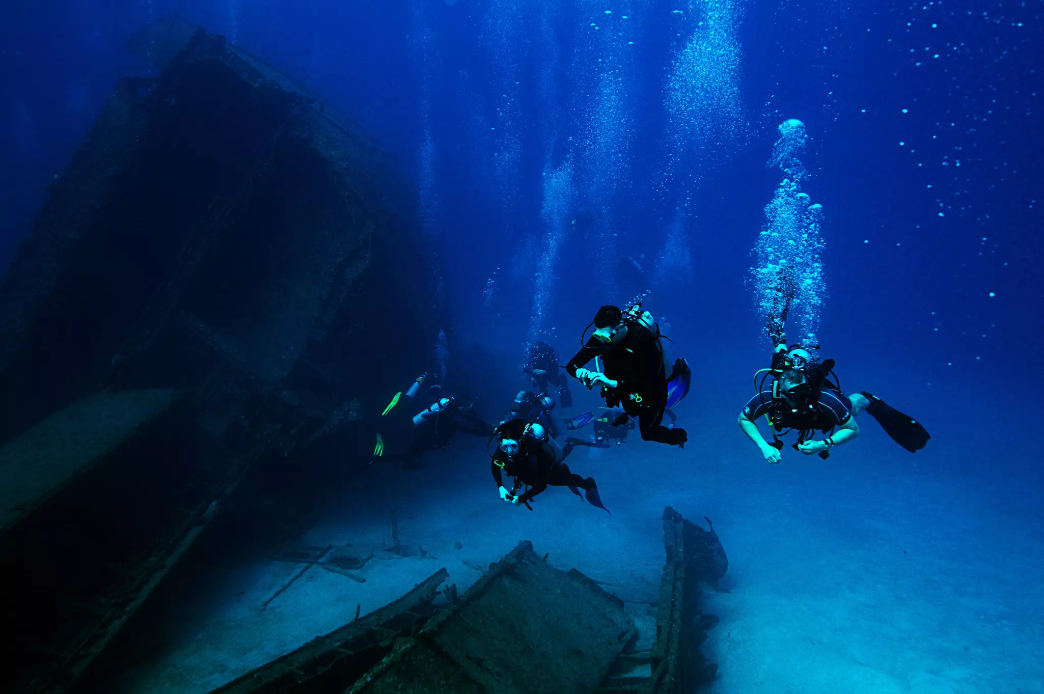 Four scuba divers descend toward the wreck of a ship on the sea floor. Bubbles stream from the divers’ masks.