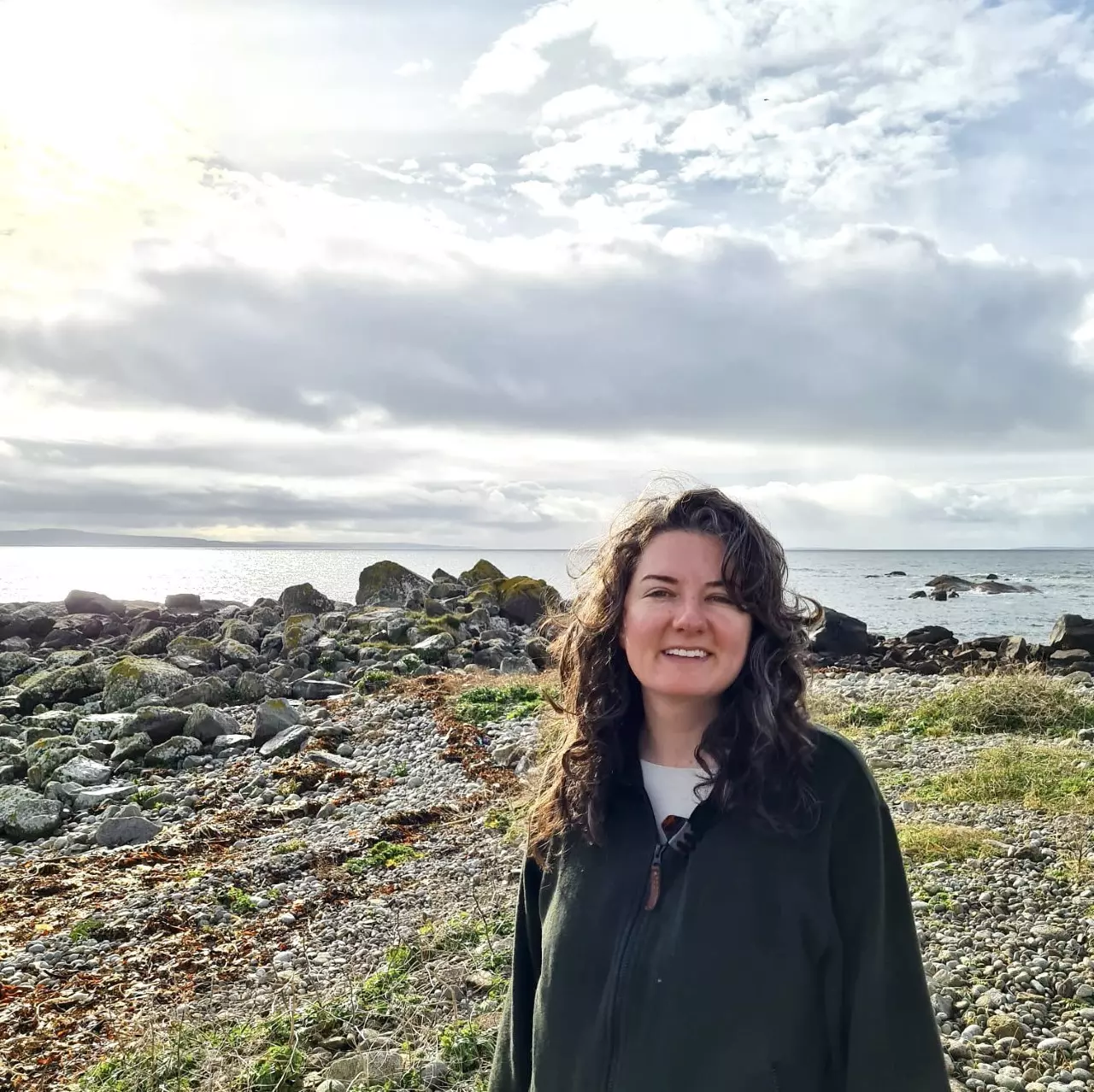 A woman wearing green stands on a rocky coastline.