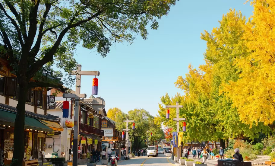 A paved street lined with bright yellow ginkgo trees, tiled-roof shops and posts with red and blue lanterns, people walking.