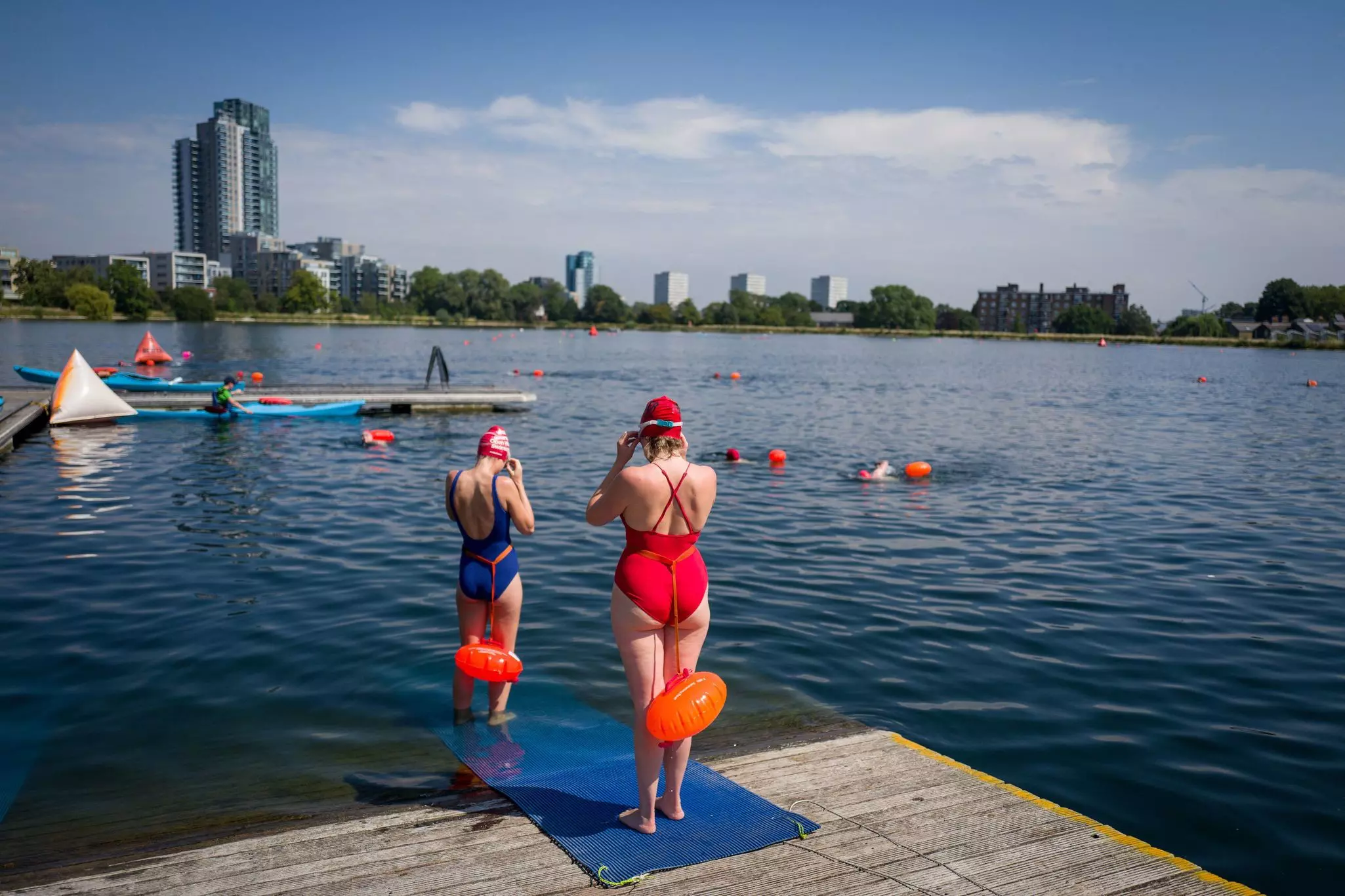 Two women with swim floats attached to their waists stand on a jetty and get ready to enter the water for a swim.