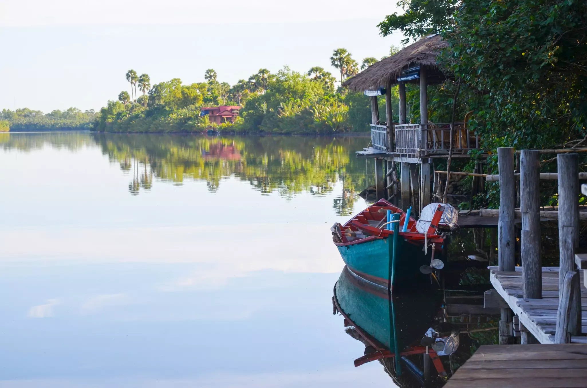 A turquoise and red boat on a placid river is pulled alongside a wooden pier; there is a home on the far shore amid dense vegetation. There are reflections on the surface of the water.