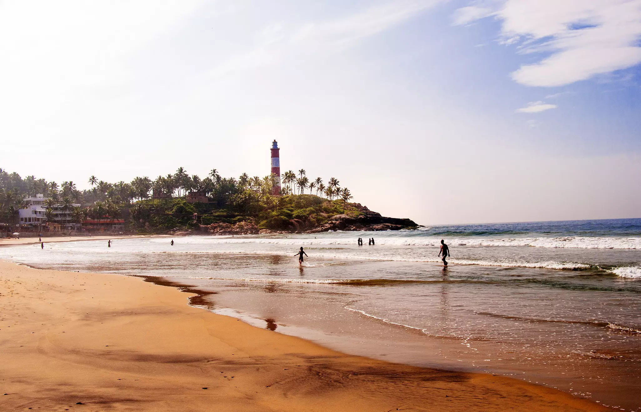 People wading into the water on a beach overlooked by a red-and-white striped lighthouse