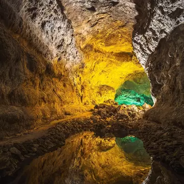 Cueva de los Verdes lava tunnel lit in yellow with a green-lit chamber ahead, its rock walls mirrored in a still pool