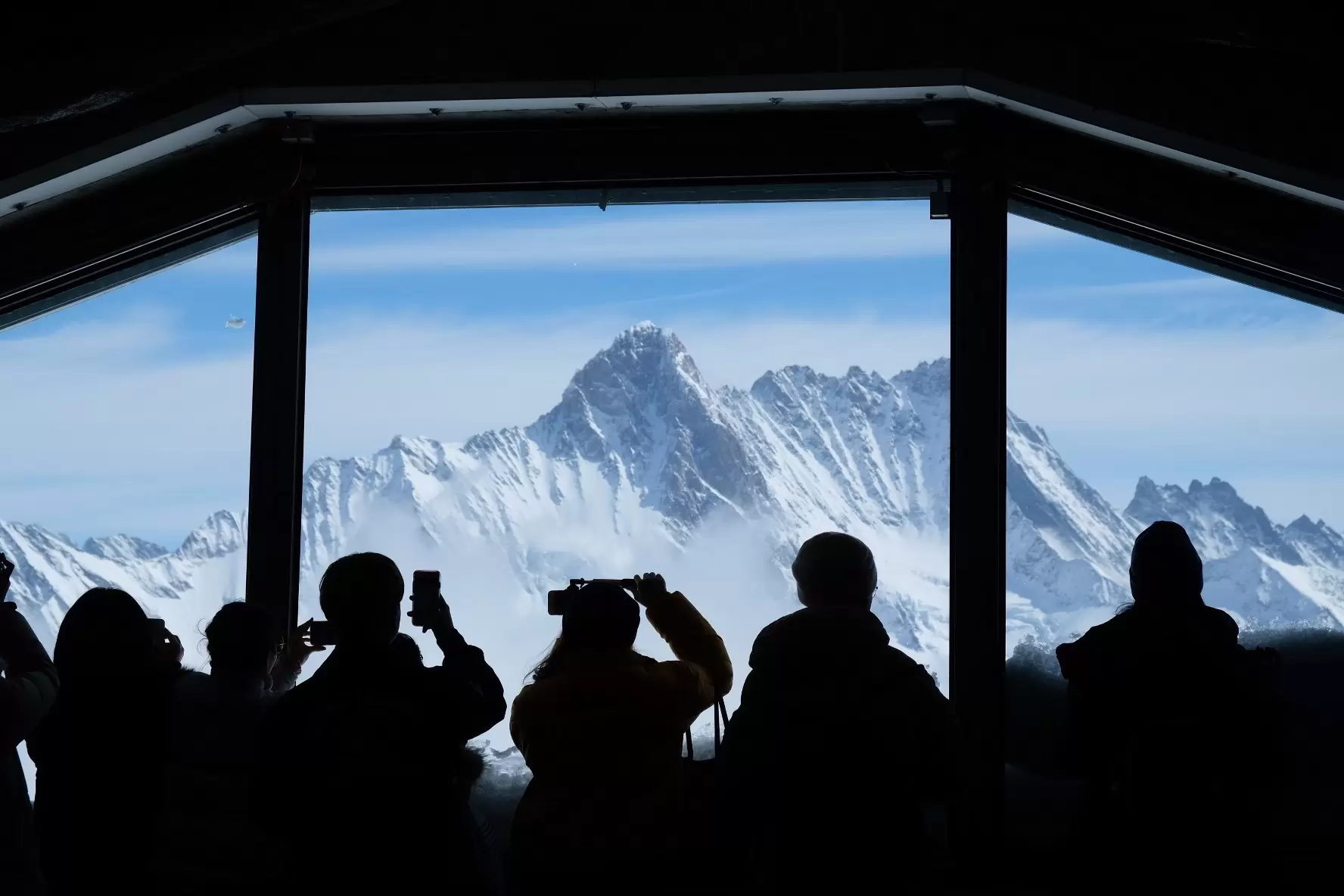 Silhouettes of tourists look out through a giant window at the snowy mountainous landscape surrounding Jungfraujoch.